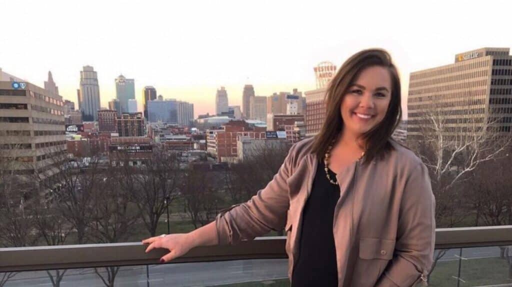 A woman smiling on a balcony with a city skyline and buildings in the background during sunset.