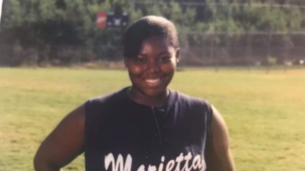 Young person in a "Marietta" sleeveless shirt smiling, standing on a grassy field with trees and fence in the background.