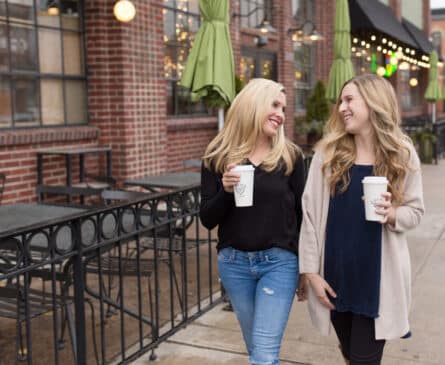 Two women walking outside, holding coffee cups, smiling and talking. They are in front of a brick cafe with green umbrellas.
