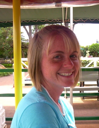 Smiling person with short blonde hair wearing a light blue shirt outdoors, seated on a bench with a park background.