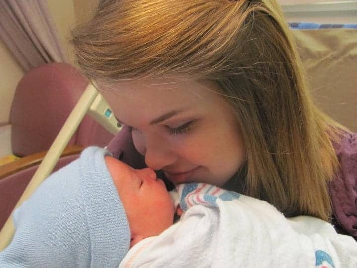A woman with blonde hair lovingly touches noses with a newborn baby wrapped in a blanket and wearing a blue hat.