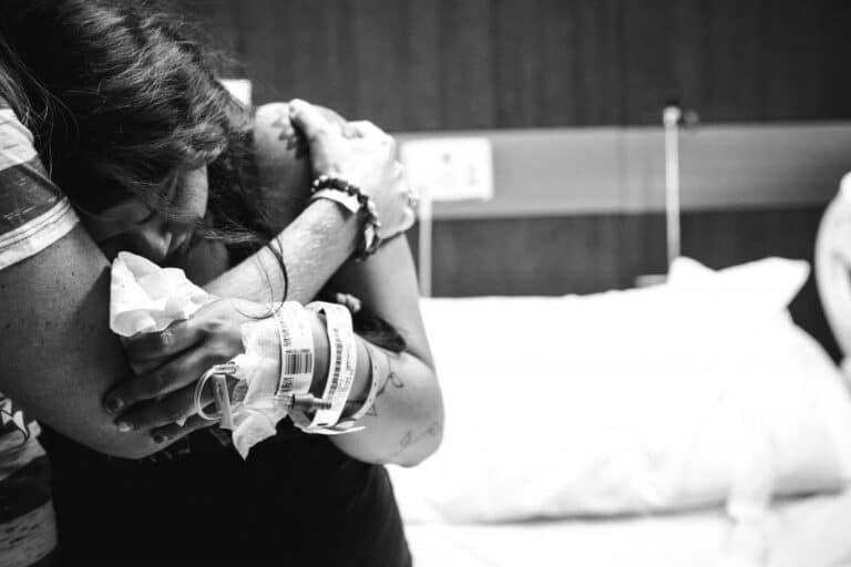 A woman with hospital wristbands is emotionally embracing someone in a hospital room, her face buried in their arms.