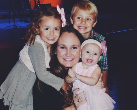 A smiling woman lovingly surrounded by three young children, one boy and two girls, all embracing each other in a warmly lit setting.