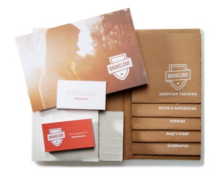 An open cardboard box displaying BraveLove materials: a photo, pamphlets, and cards about adoption with words like 