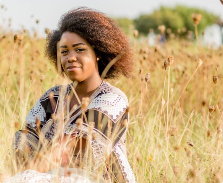 A person with curly hair, wearing a patterned top, sitting in a field of dry plants, smiles softly with trees in the background.