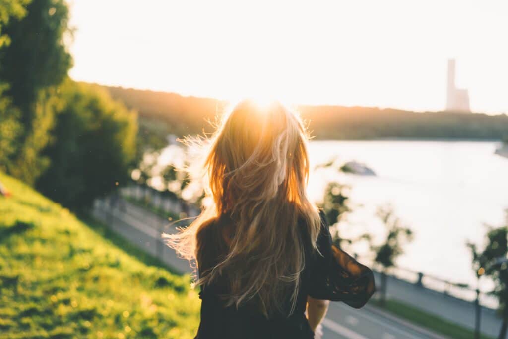 Woman with long hair stands on a grassy hill overlooking a river and cityscape at sunset, with the sun creating a bright glow behind her.