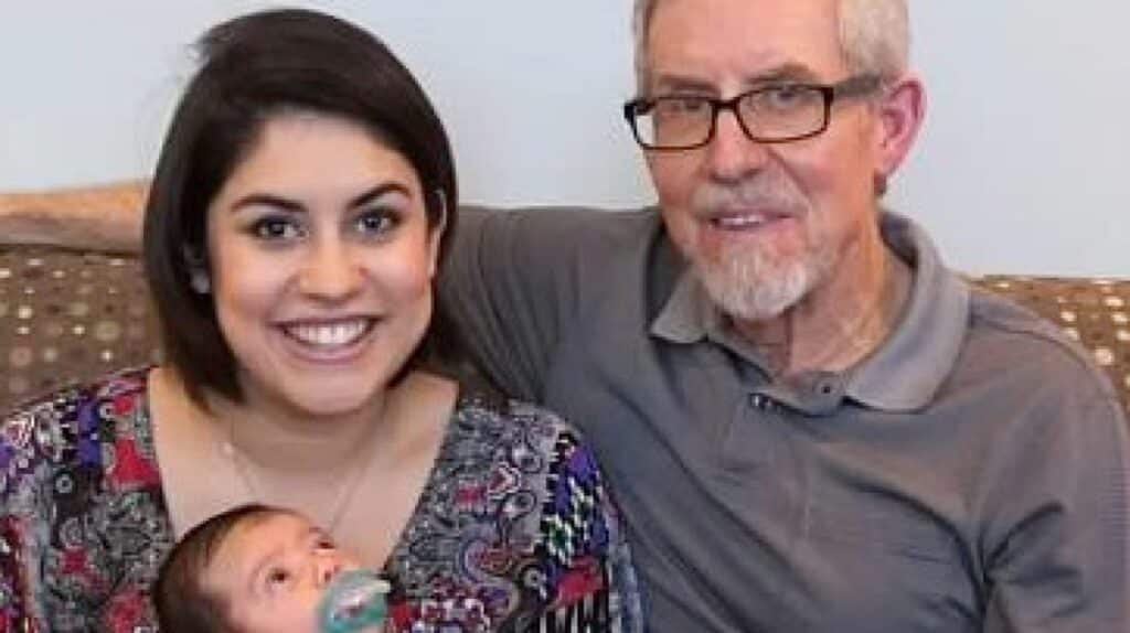 A woman holding a baby bottle-feeding an infant is seated next to an older man with glasses and gray hair on a couch.