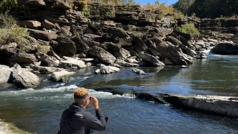 Person sitting by a rocky riverbank, looking at the water with rocks and greenery in the background.