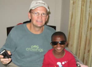 Man in a cap and a child wearing sunglasses smile while sitting together indoors near a curtain.