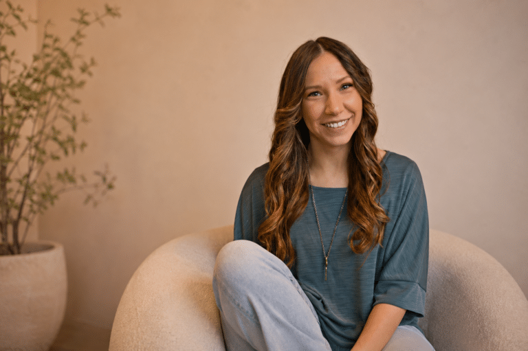 Smiling woman with long brown hair sits on a round beige chair near a potted plant, wearing a teal shirt and jeans.