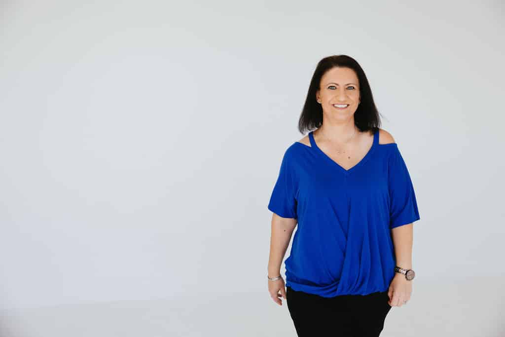 Woman with straight dark hair wearing a bright blue top and black pants stands smiling against a plain white background.