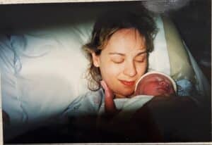 A woman lies in bed smiling with her eyes closed, holding a newborn baby wrapped in a blanket close to her.