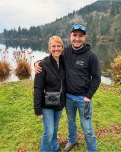 Smiling woman and man standing together by a lake, with trees and hills in the background on a cloudy day.