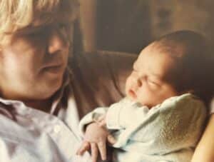 An adult with blond hair gently holds a sleeping newborn baby wrapped in a light blanket.