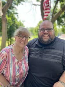 Two people smiling outdoors, standing close together, with trees and a blurred American flag in the background.