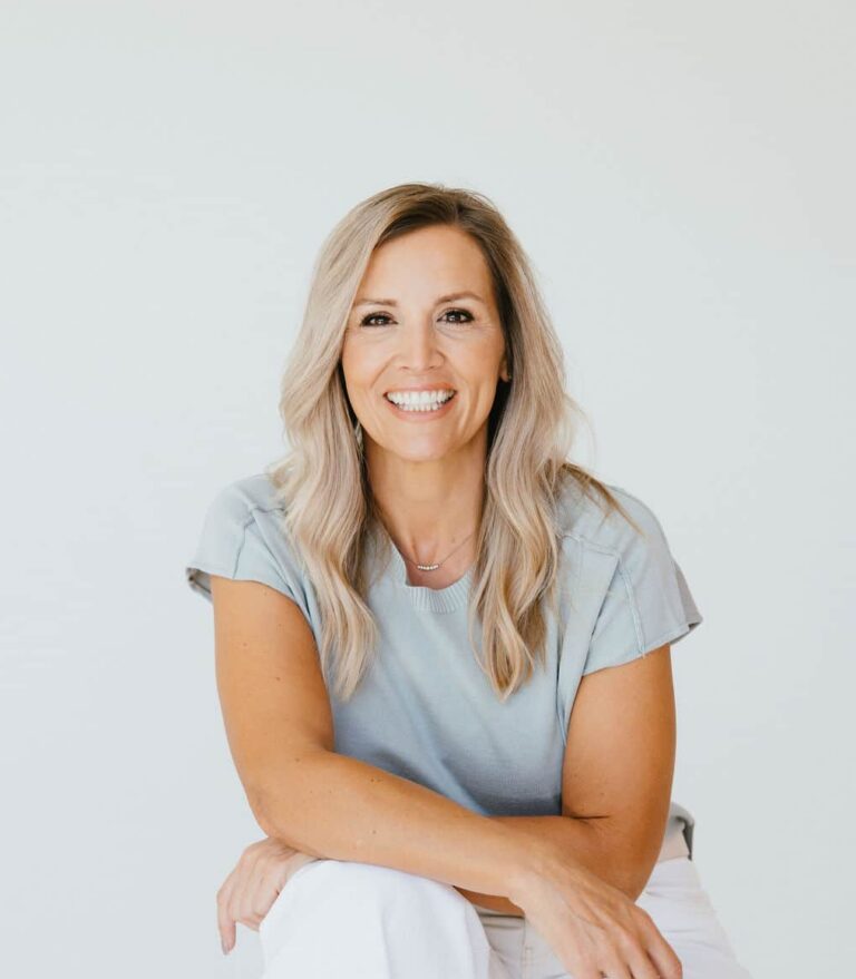 Smiling woman with long blonde hair, wearing a light blue shirt and white pants, seated against a plain white background.
