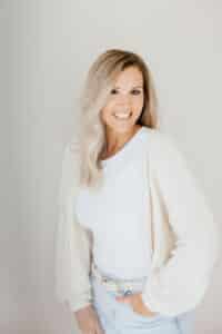 Smiling woman with long blonde hair wearing a white shirt, light cardigan, and jeans, standing against a neutral background.