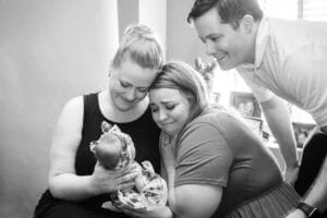 Three adults smile and cry as they lovingly gaze at a newborn baby held by one of the women, in a cozy room.