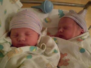 Two newborn babies in striped hats are swaddled in blankets and sleeping side by side in a hospital setting.