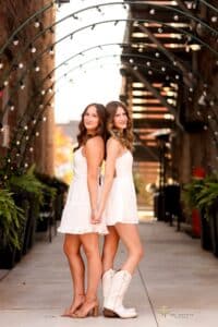 Two young women in white dresses stand back-to-back and hold hands, smiling under an arched walkway decorated with string lights.