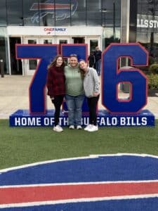 Three people stand smiling in front of a large “716” sign at the Buffalo Bills stadium entrance.