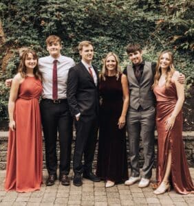 Six people pose outdoors in formal attire, standing on a stone walkway with greenery behind them.
