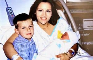 A woman in a hospital bed holds a newborn and smiles with a young boy, likely her son, sitting close beside her.