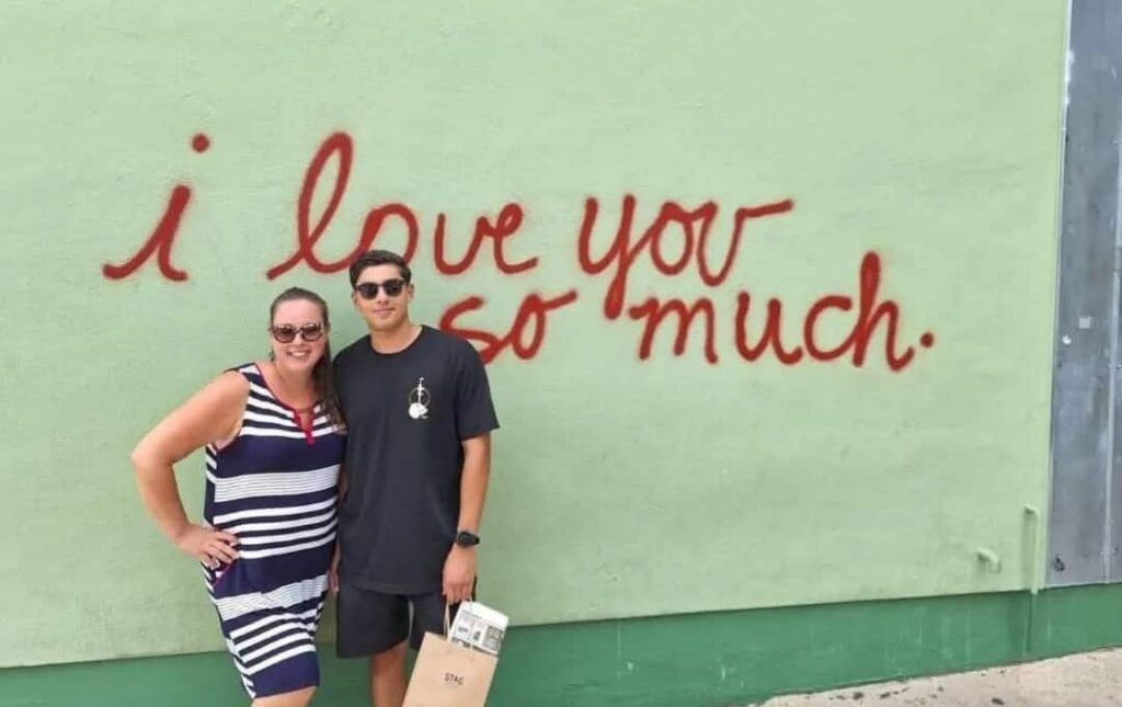 A woman and a young man posing in front of a green wall with the red spray-painted text “i love you so much.”